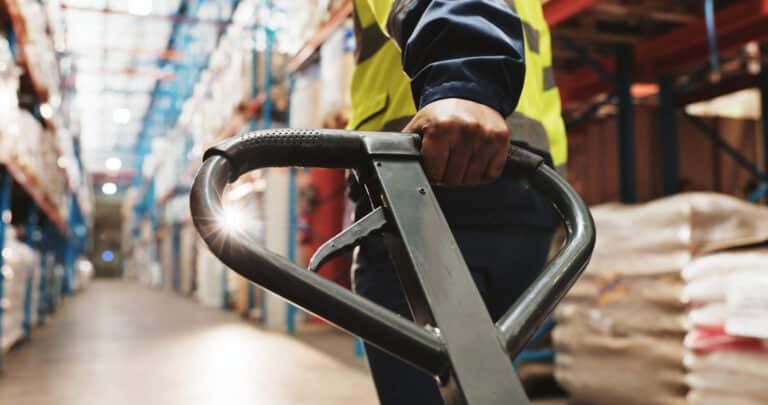 Warehouse worker gripping a pallet-truck handle between storage shelves.